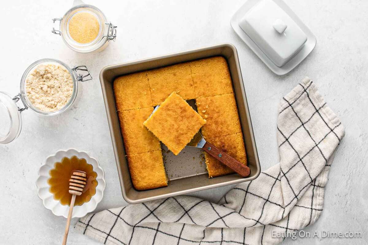 A square pan of freshly baked gluten-free cornbread with one piece being lifted out by a spatula. Surrounding the pan are a jar of cornmeal, a honey dipper on a small plate, and a kitchen towel.