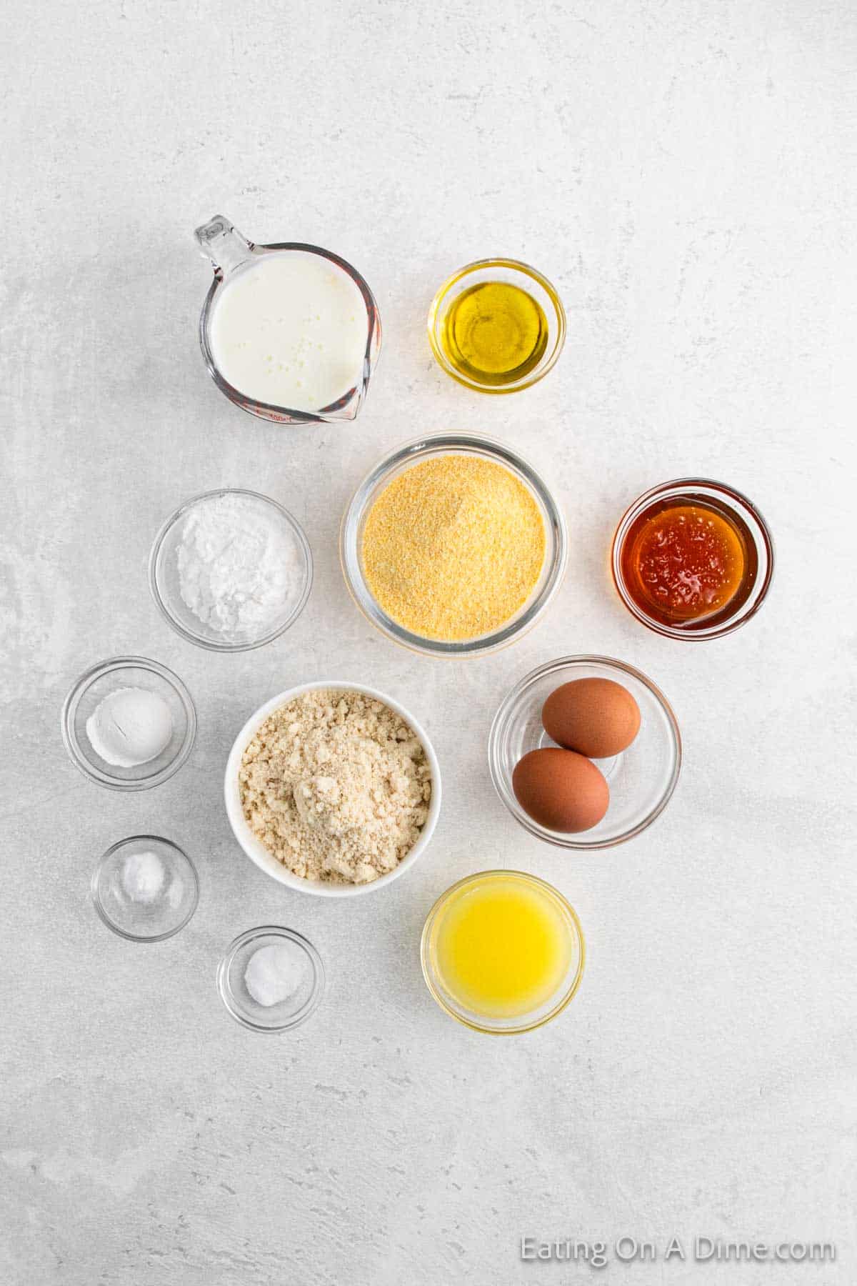 Top-down view of cornbread baking ingredients arranged on a white surface, including a pitcher of milk, bowls of gluten-free cornmeal, flour, sugar, salt, baking powder, oil, melted butter, honey, and two brown eggs.