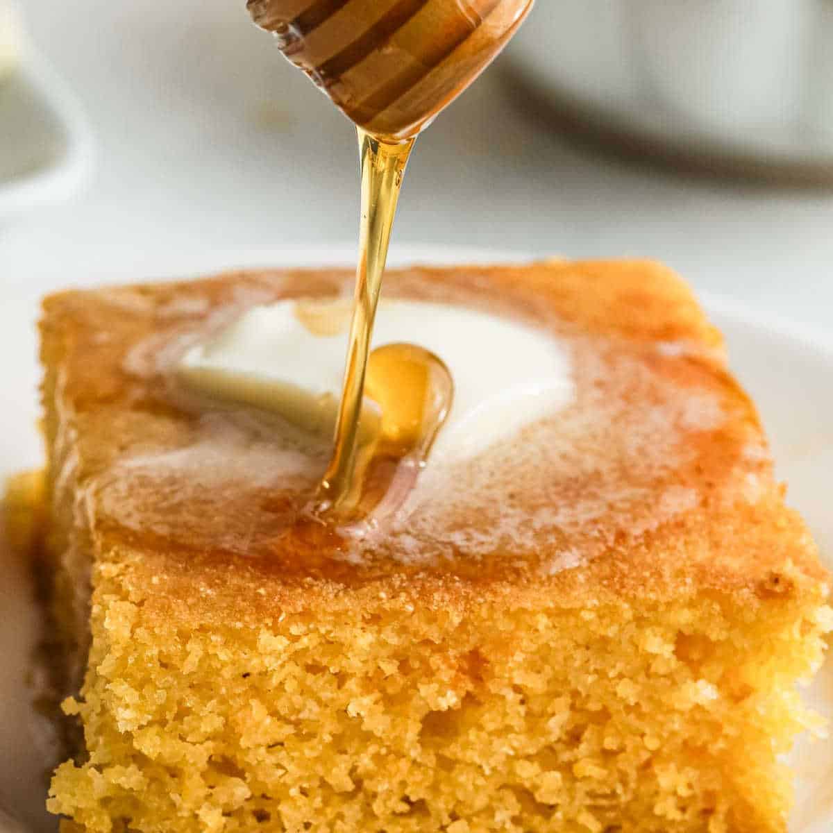 A close-up of a slice of gluten-free cornbread on a plate, topped with a small pat of butter. Honey is being drizzled from a wooden honey dipper onto the cornbread, partially covering the butter.