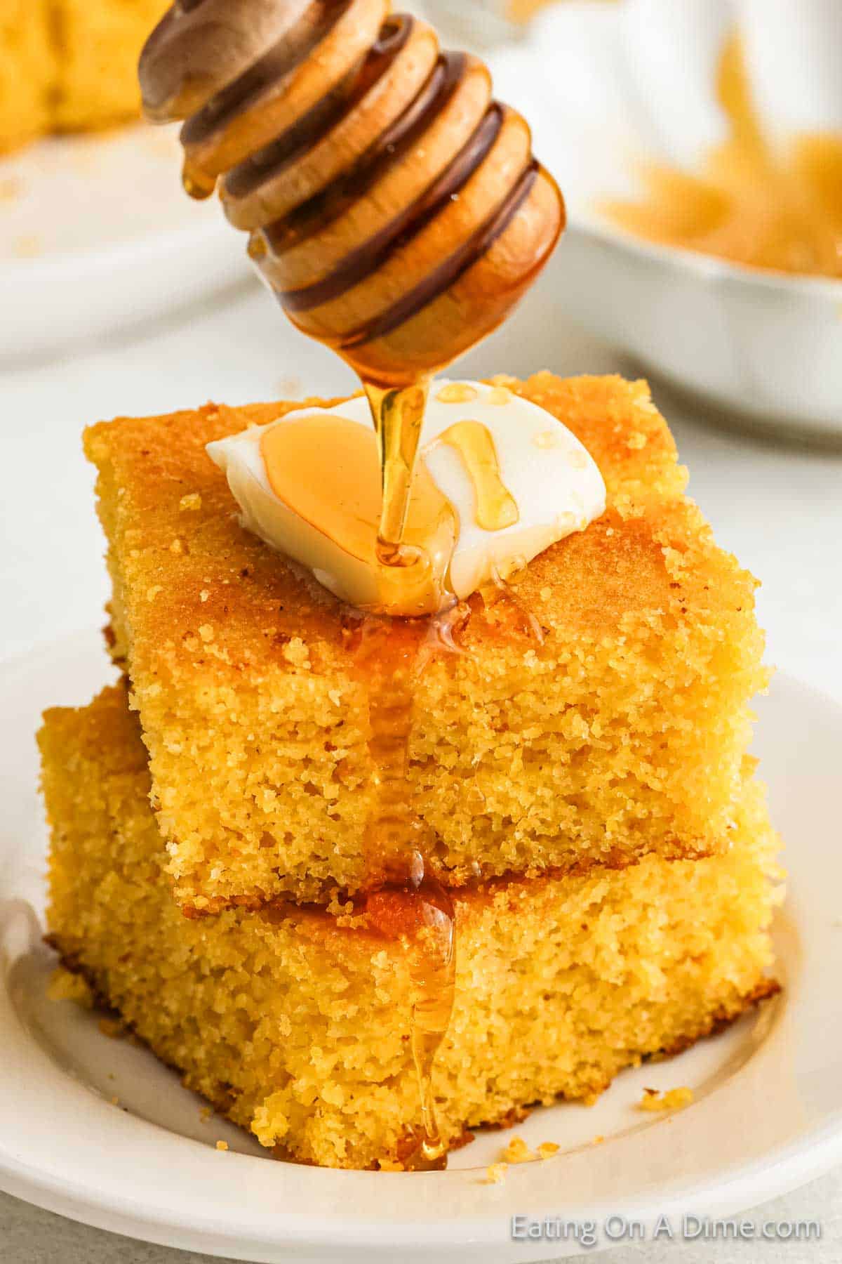 Two stacked pieces of golden, gluten-free cornbread sit on a white plate, crowned with a pat of butter. Honey drizzles from a wooden dipper onto the butter, creating a glossy stream over the delicious cornbread.