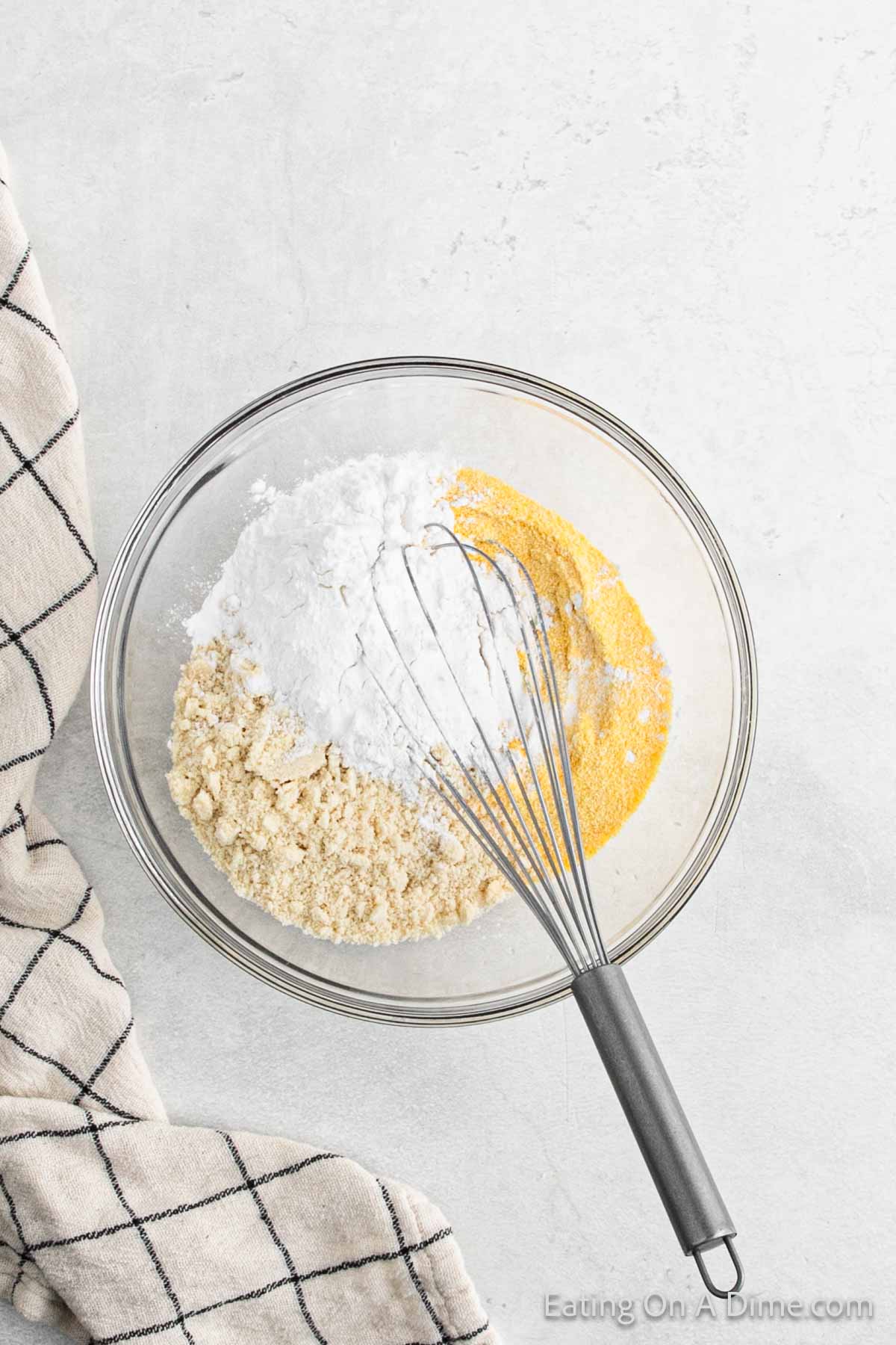 A glass bowl filled with cornmeal, flour, and baking powder, ready to make delicious cornbread. A whisk is poised inside, and a checkered cloth peeks from the left side of the image.