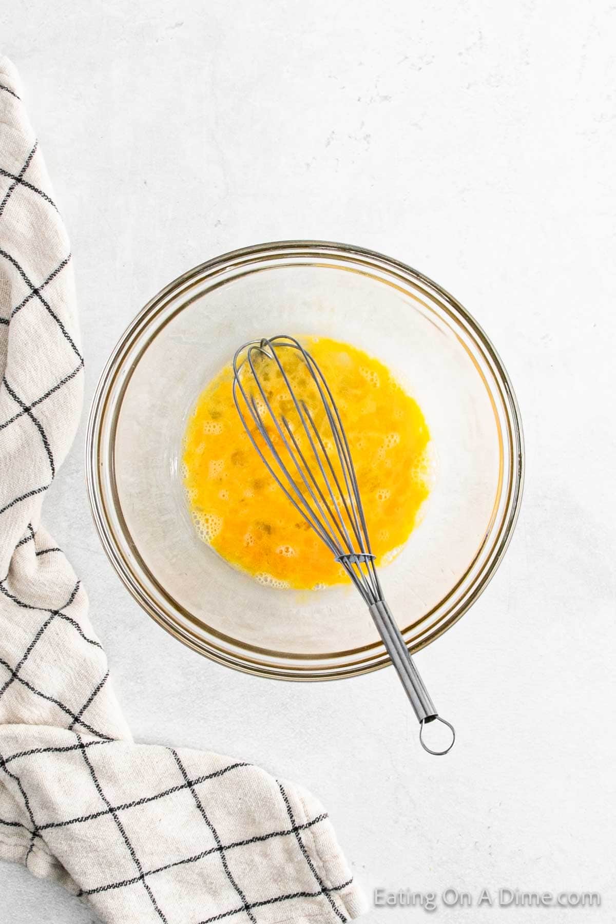 A clear glass bowl containing beaten eggs with a metal whisk resting inside awaits the next step in making gluten-free cornbread. A white dish towel with black grid lines is draped on the left. The background is a light, textured surface.