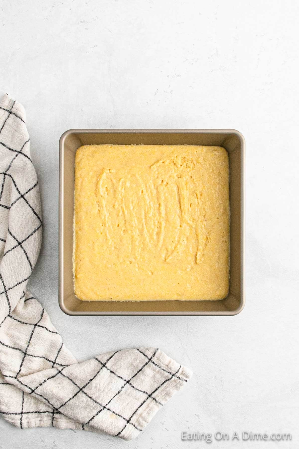 A square baking pan filled with freshly baked, gluten-free cornbread rests on a light-colored surface. A black and white checkered kitchen towel is draped to the side, complementing the simple and minimalistic background.