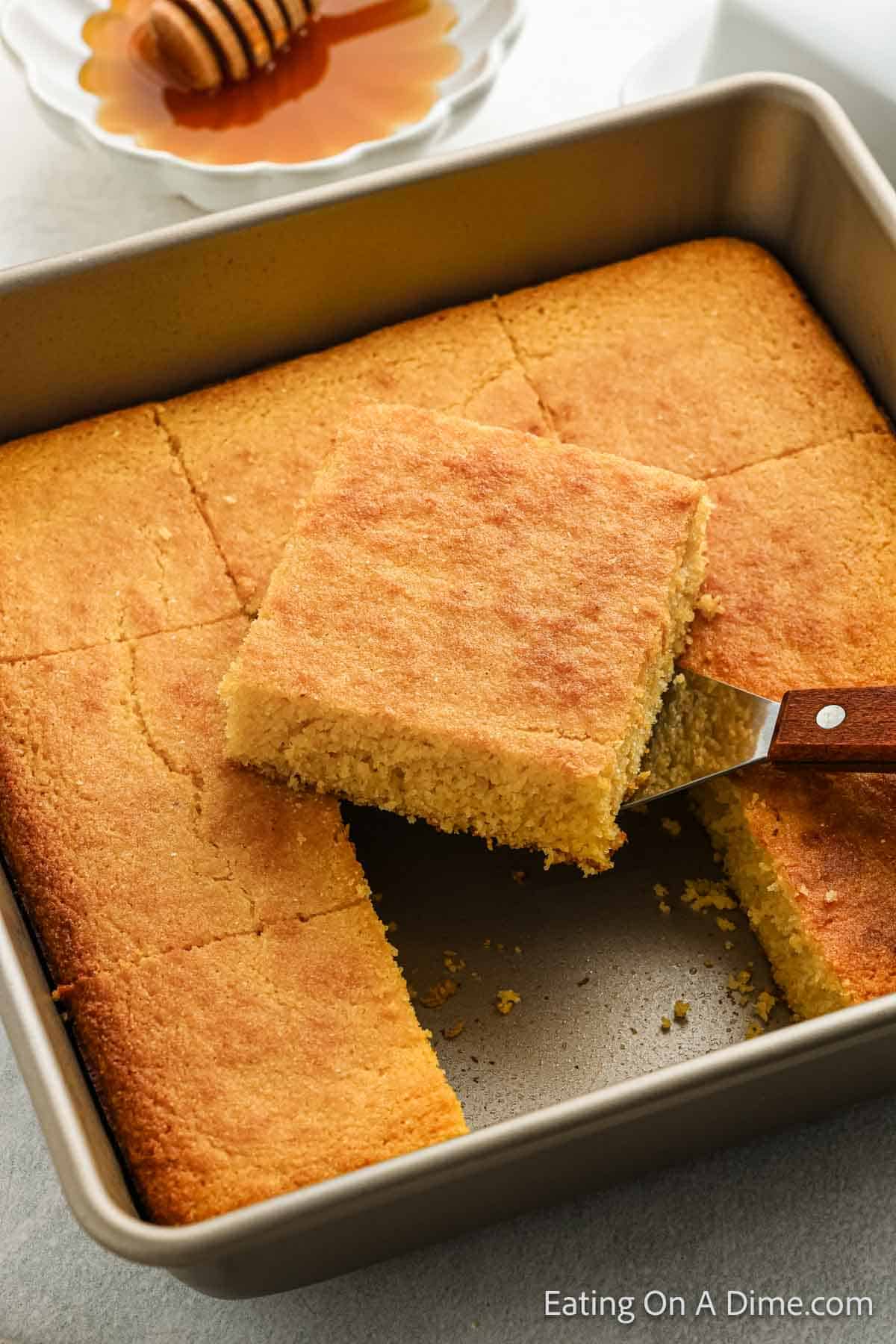 A square pan holds freshly baked gluten-free cornbread, cut into perfect squares. One square is being lifted out with a spatula, revealing its golden goodness. In the background, there's a small dish of honey with a honey dipper ready to add a sweet touch.
