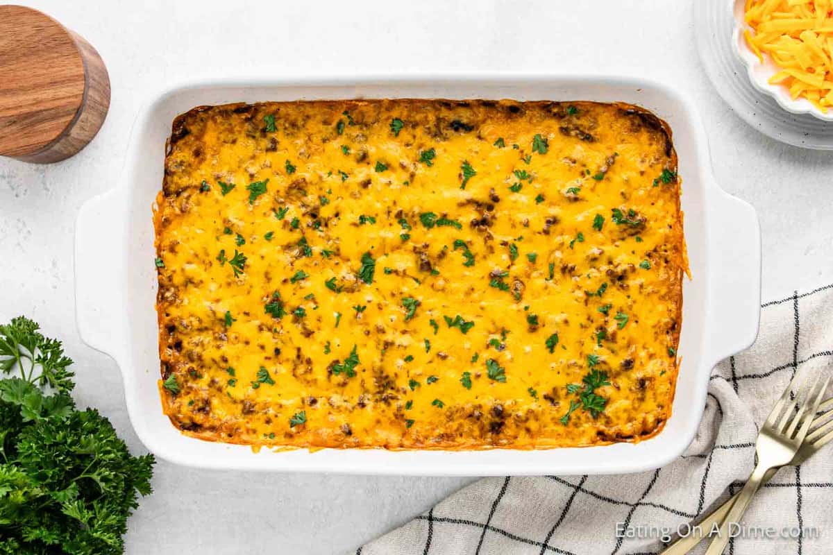 A rice and hamburger casserole bakes beautifully in a white rectangular dish, topped with melted cheese and garnished with parsley. It's elegantly set on a light surface beside a wooden lid, fresh parsley, a striped cloth, and a bowl of shredded cheese.