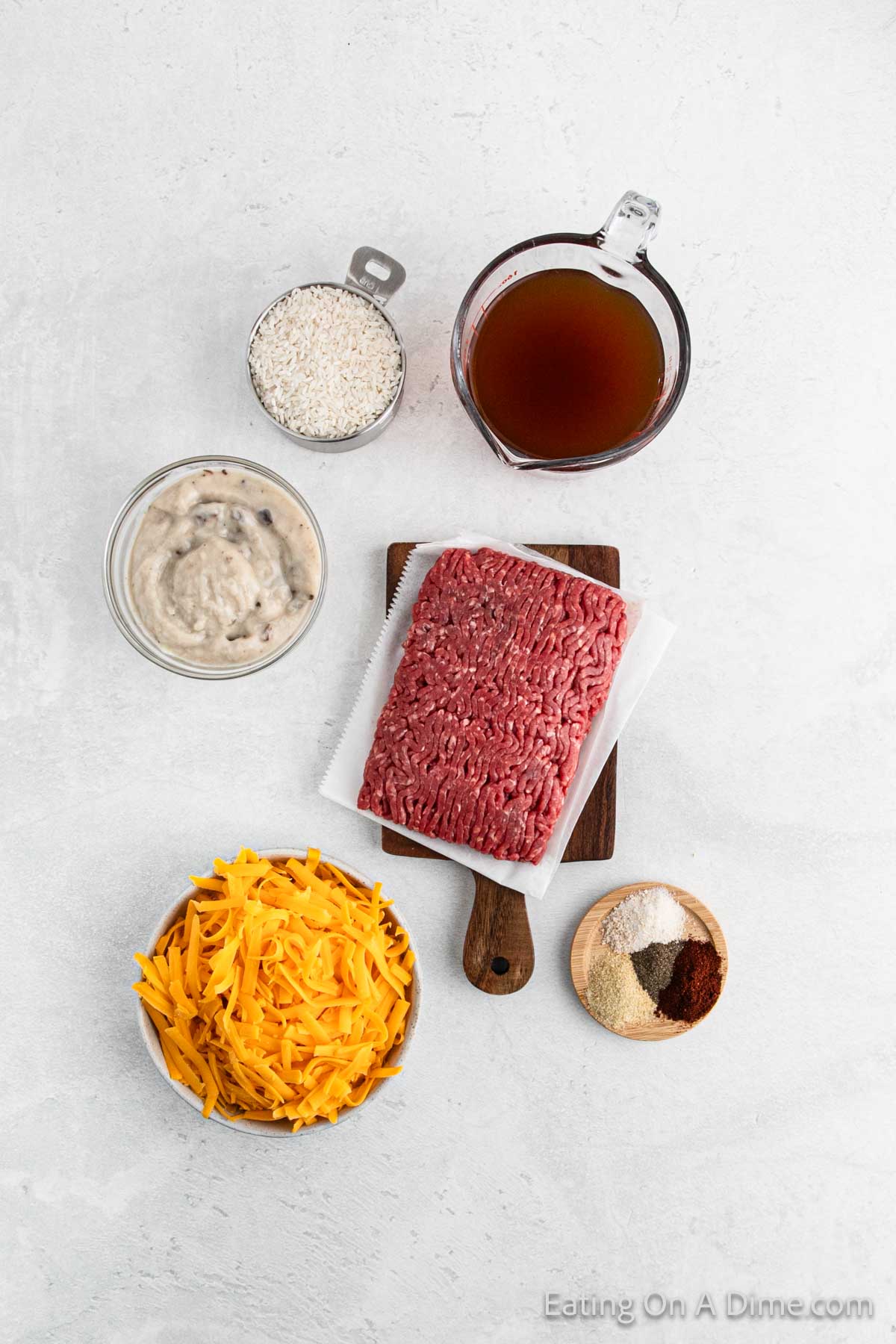 Top view of casserole ingredients for a delicious recipe, featuring ground beef, shredded cheese, rice, broth, cream soup, and spices on a light surface.