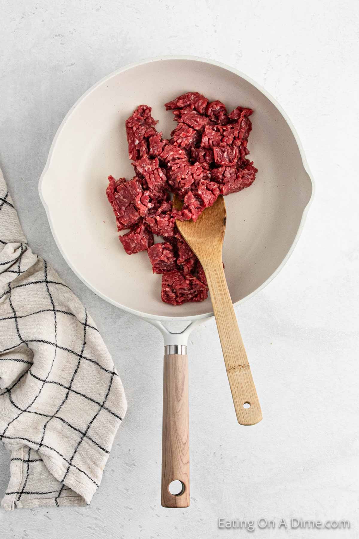 A frying pan with raw ground beef chunks, reminiscent of a hearty hamburger, and a wooden spatula rests on a light surface. A checkered kitchen towel lies beside the pan, setting the stage for a comforting meal.