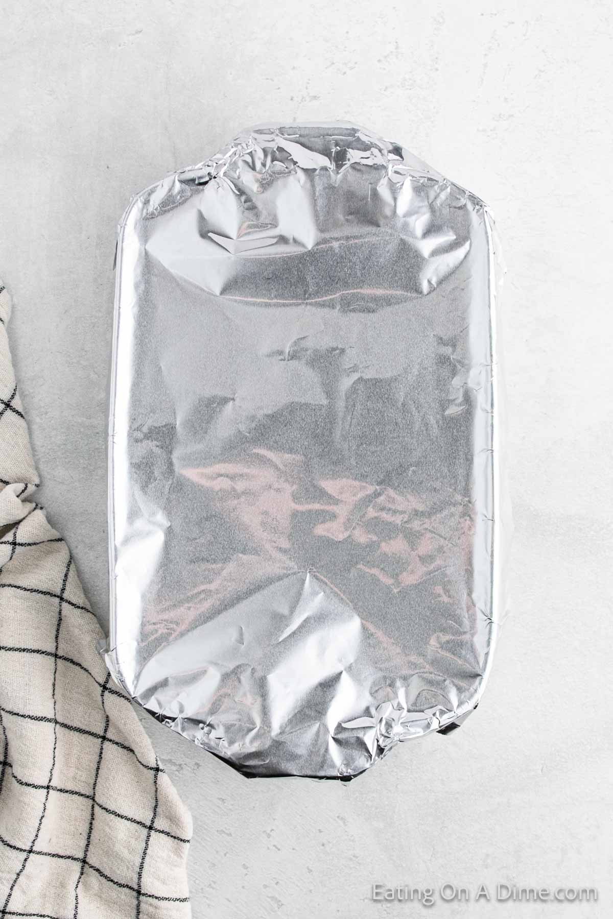 A baking dish covered with aluminum foil rests on a light gray surface, hinting at the steaming casserole inside. A black and white checkered cloth is partially visible on the left side, adding a touch of cozy charm to the setting.