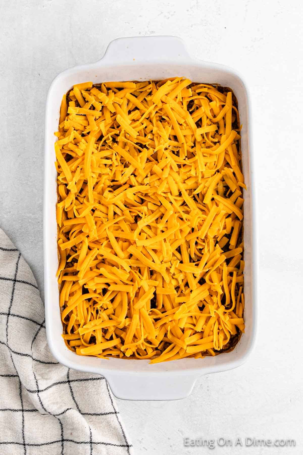 A casserole dish brimming with shredded cheddar cheese and savory hamburger sits on a textured surface. A black and white checkered cloth is partially visible on the left side, adding a touch of charm to this hearty delight.