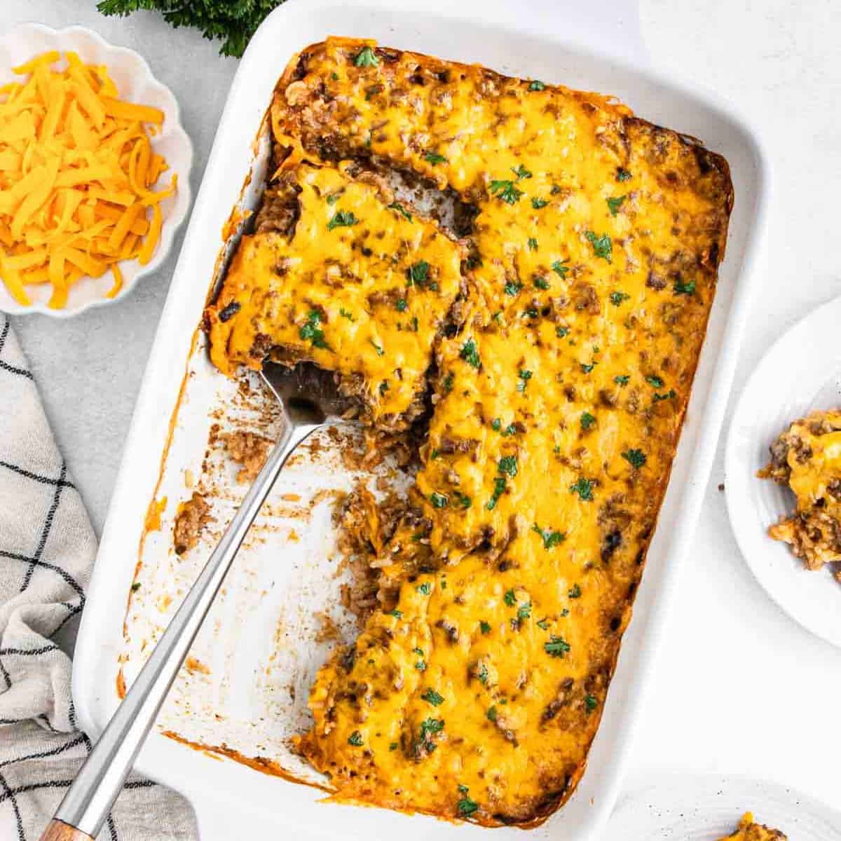 A casserole dish brimming with cheesy ground beef and noodle casserole, topped with melted cheddar and fresh parsley. A serving spoon rests inside, next to a square piece on a white plate. Shredded cheese and a cloth lie beside the hearty hamburger delight.