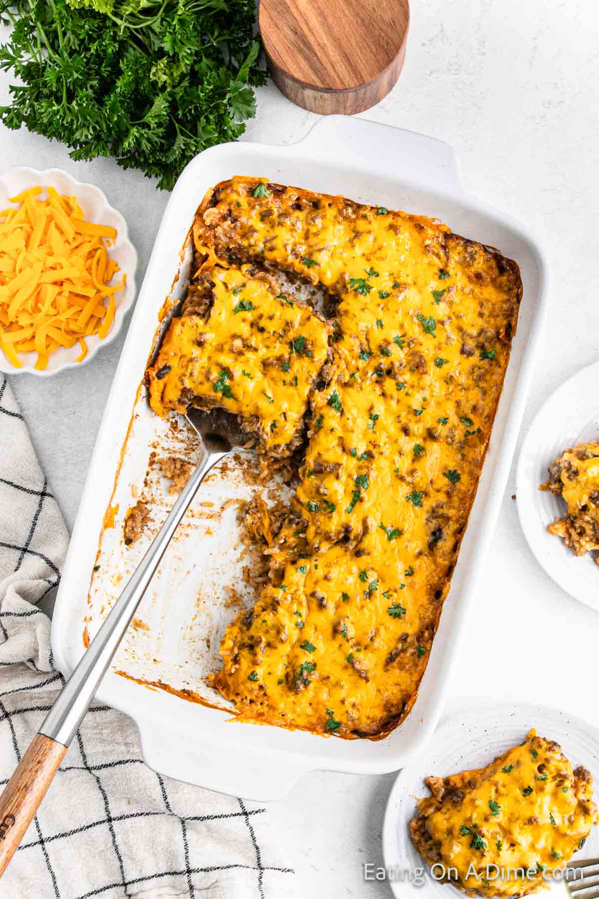 A casserole dish brimming with a savory beef, rice, and cheese medley is topped with melted cheddar and garnished with herbs. A serving rests enticingly on a nearby plate, while in the background, a bowl of shredded cheddar and fresh parsley awaits.