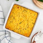 A square baking dish filled with golden-brown Mexican Cornbread, slightly cracked on top, sits on a white surface next to a striped towel and a serving utensil.