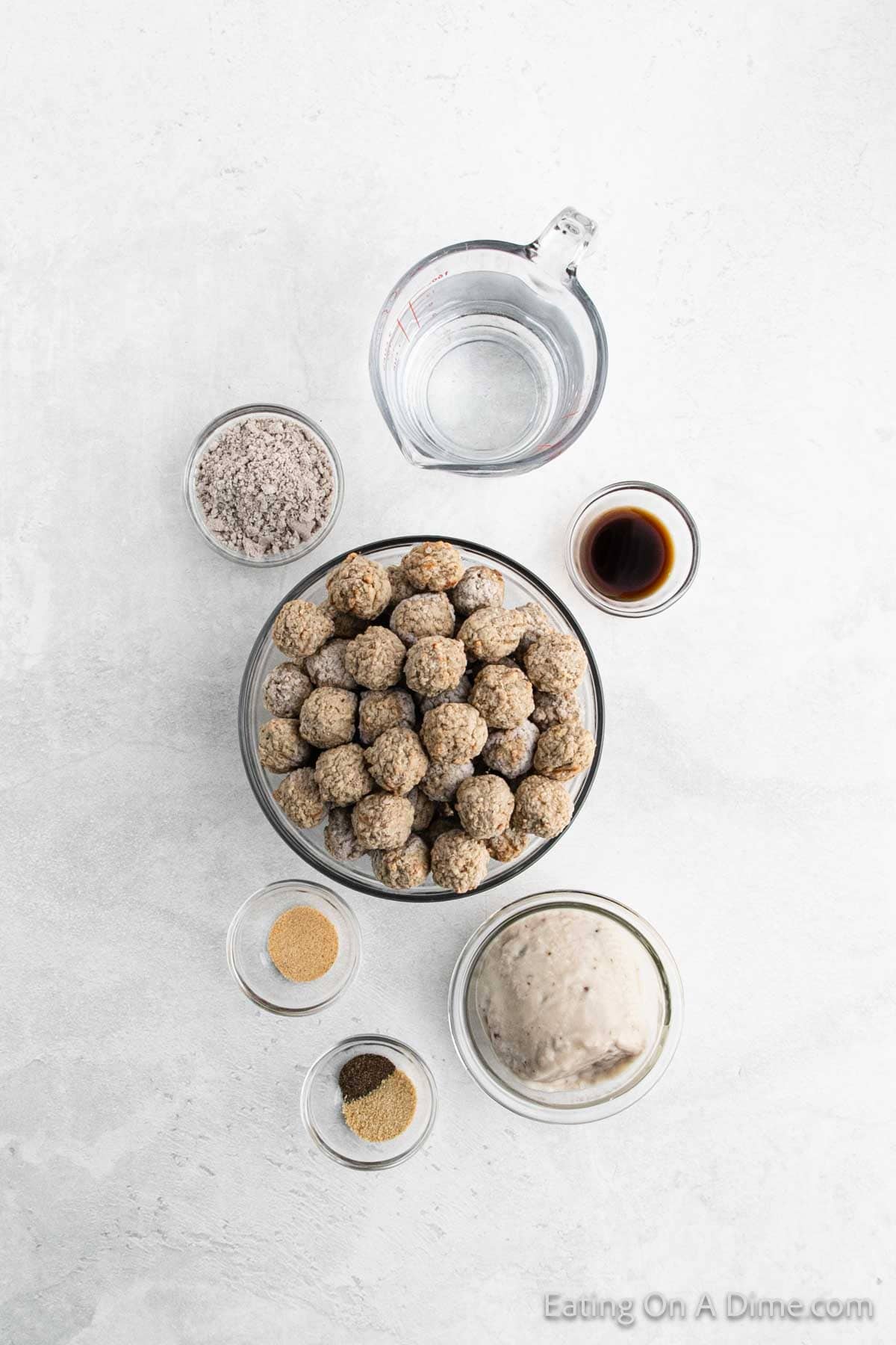 Ingredients for Swedish meatballs on a white surface include a bowl of raw meatballs, a measuring cup of water, soy sauce, flour, cream, and small bowls of spices. Perfect for simmering in a slow cooker until tender with rich gravy.