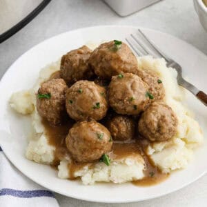 A plate of mashed potatoes topped with slow cooker meatballs drizzled in rich brown gravy. The dish is garnished with chopped herbs. A fork is placed beside the plate on a light-colored tablecloth.