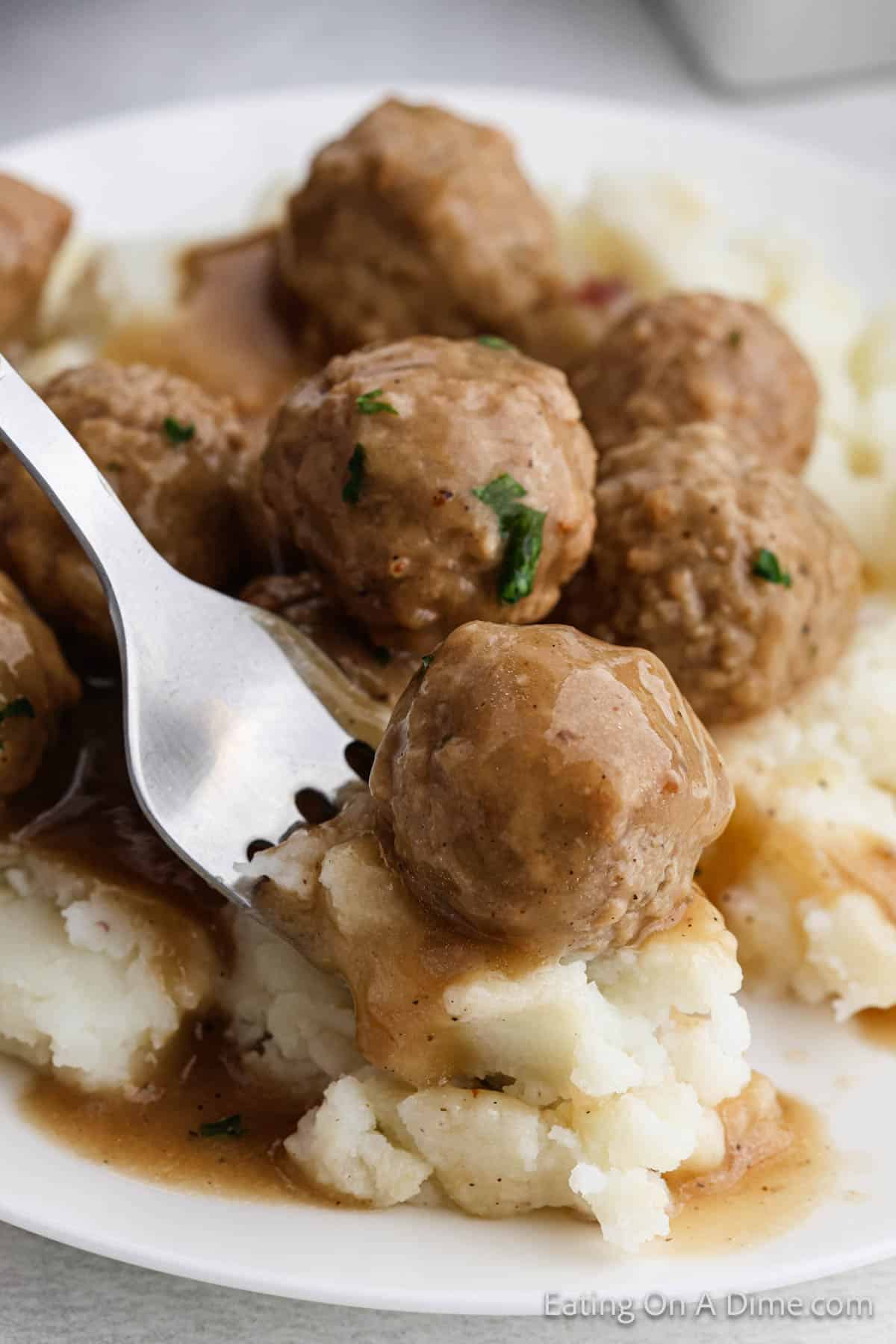 A fork holding a slow-cooked meatball sits on a plate of creamy mashed potatoes, topped with rich gravy. Several meatballs are on the plate, garnished with fresh herbs.