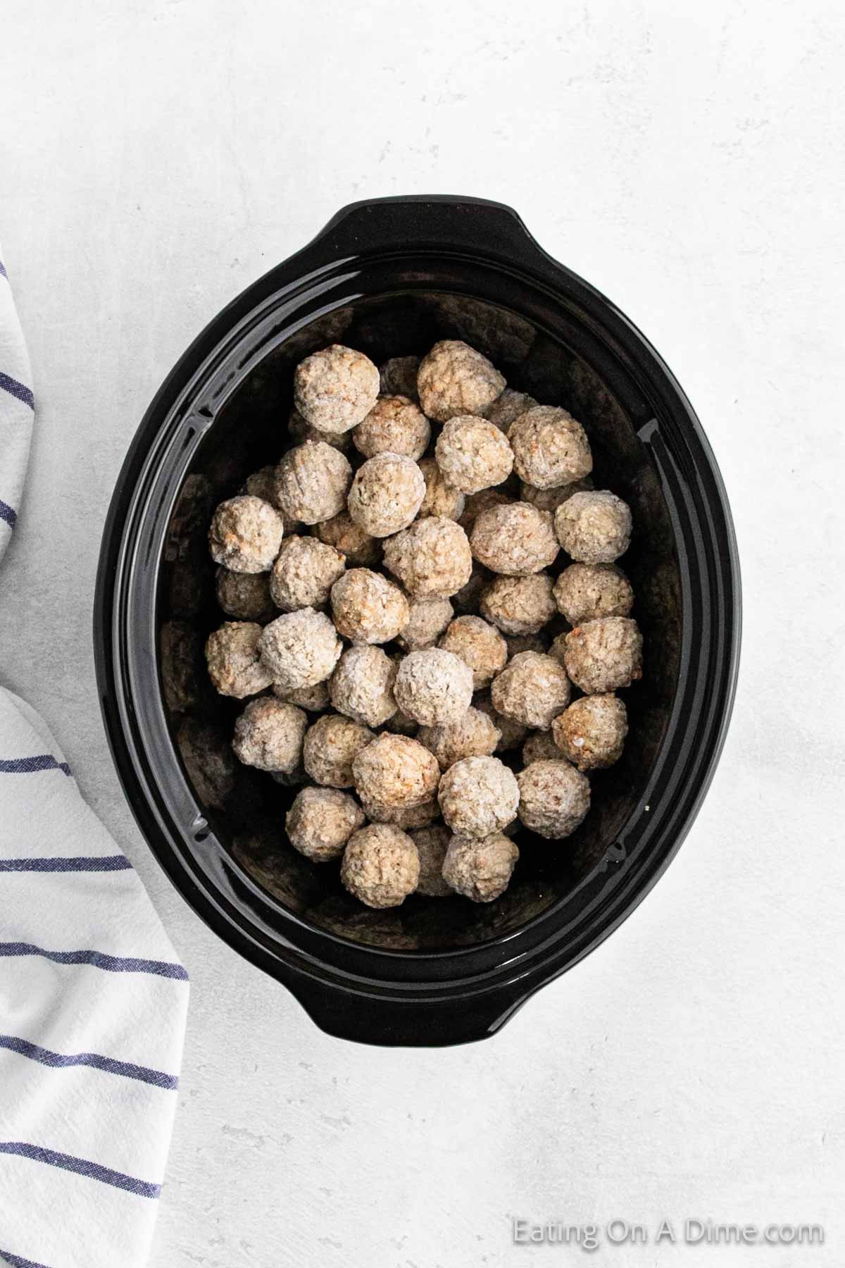 A black slow cooker brimming with round, seasoned meatballs bathed in rich gravy sits elegantly on a light gray surface. Beside it, a blue and white striped cloth adds a touch of charm to the scene.