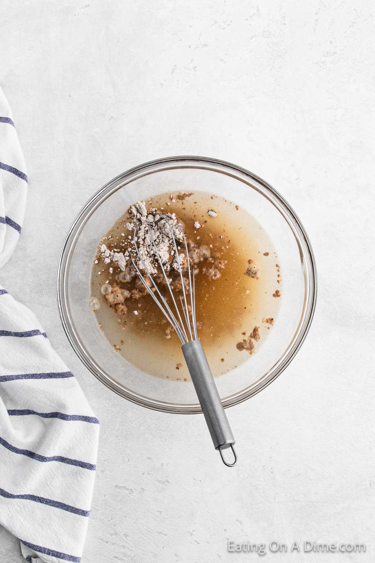A glass bowl contains a mixture of liquid and clumped brown sugar, being whisked into a savory gravy. A metal whisk is placed inside the bowl. A striped kitchen towel is partially visible on the left side atop a light textured surface.