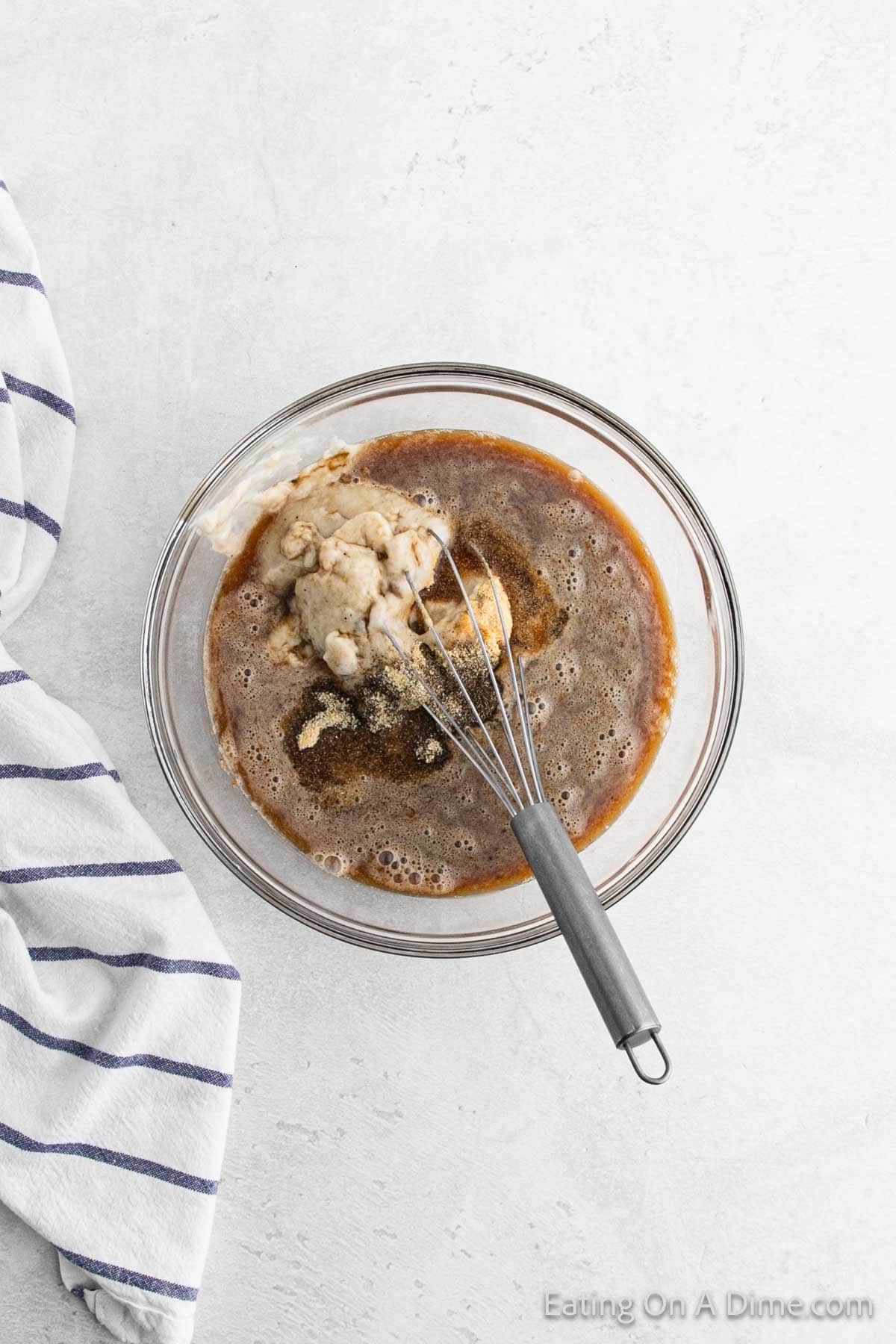 A glass bowl filled with a rich, brown gravy mixture and a clump of dough, with a whisk resting inside. The bowl is on a light gray surface next to a striped kitchen towel, ready to meet hearty meatballs for an irresistible dish.