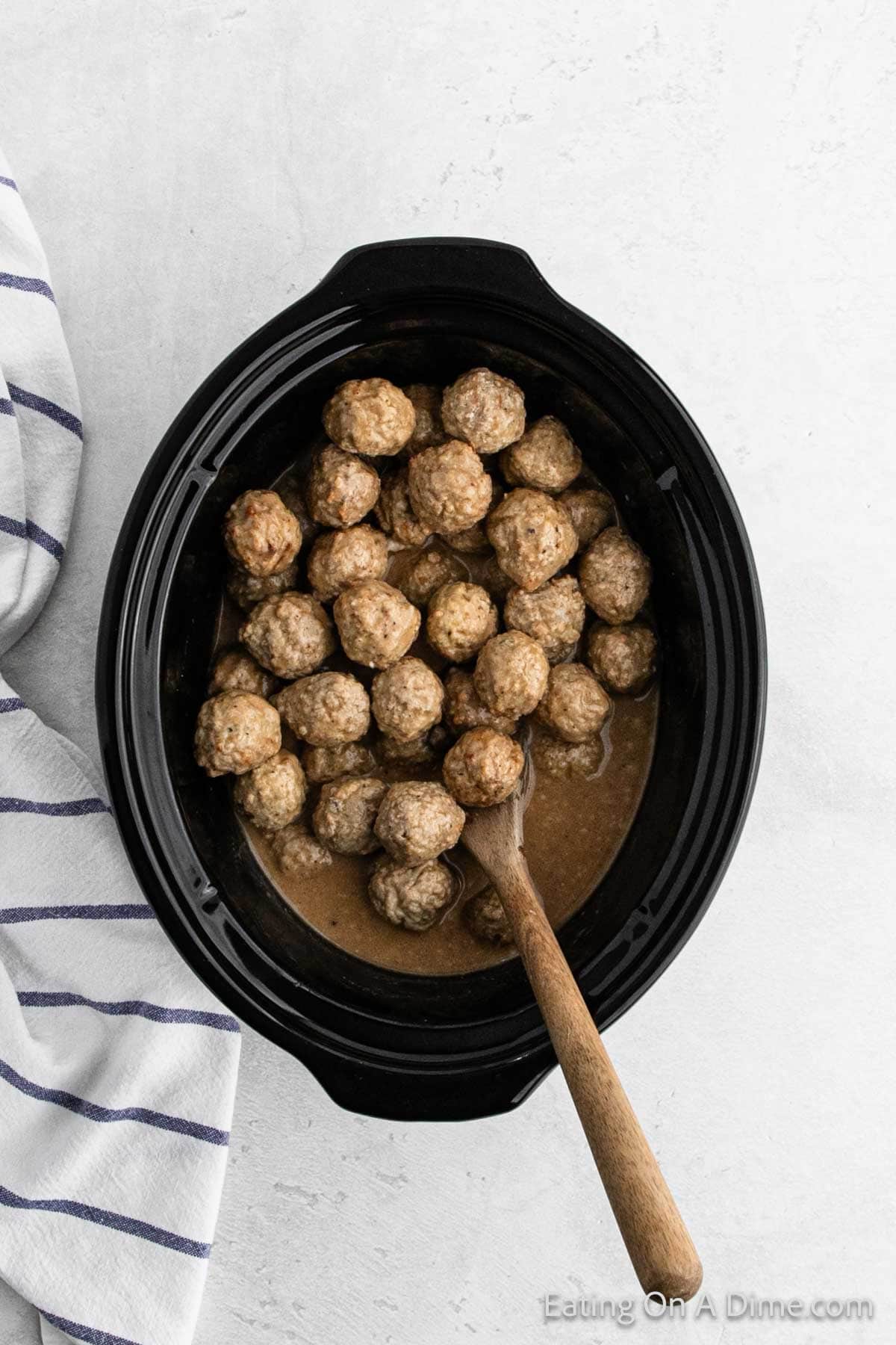 A black slow cooker brims with savory meatballs bathed in rich gravy, a wooden spoon poised at the side. To the left, a white and blue striped cloth lies gracefully on a light-colored surface.