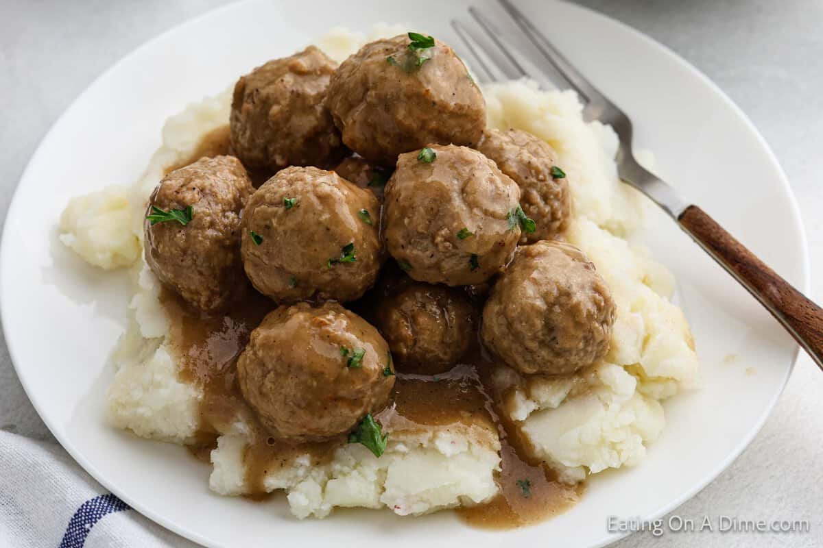 A white plate holds mashed potatoes crowned with slow-cooked meatballs, all smothered in rich brown gravy and garnished with chopped parsley. A fork rests beside the dish, inviting a savory bite.