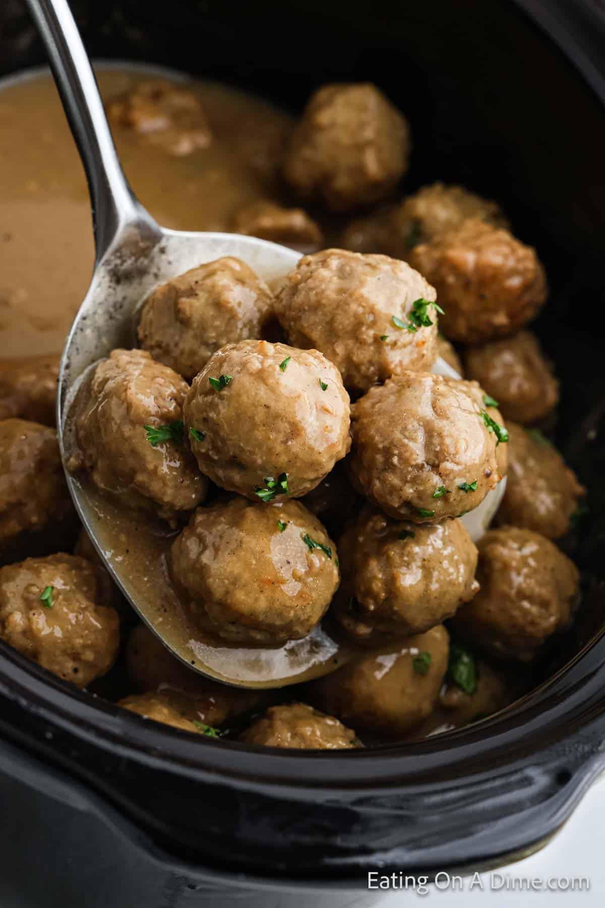 A close-up of a spoon lifting several meatballs drenched in savory brown gravy from a black slow cooker. The meatballs, sprinkled with parsley bits, are nestled among more delicious meatballs and rich sauce below.