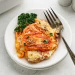A serving of cheesy tomato casserole and Sweet Potato Au Gratin, garnished with fresh parsley, is plated with a fork on a white dish.