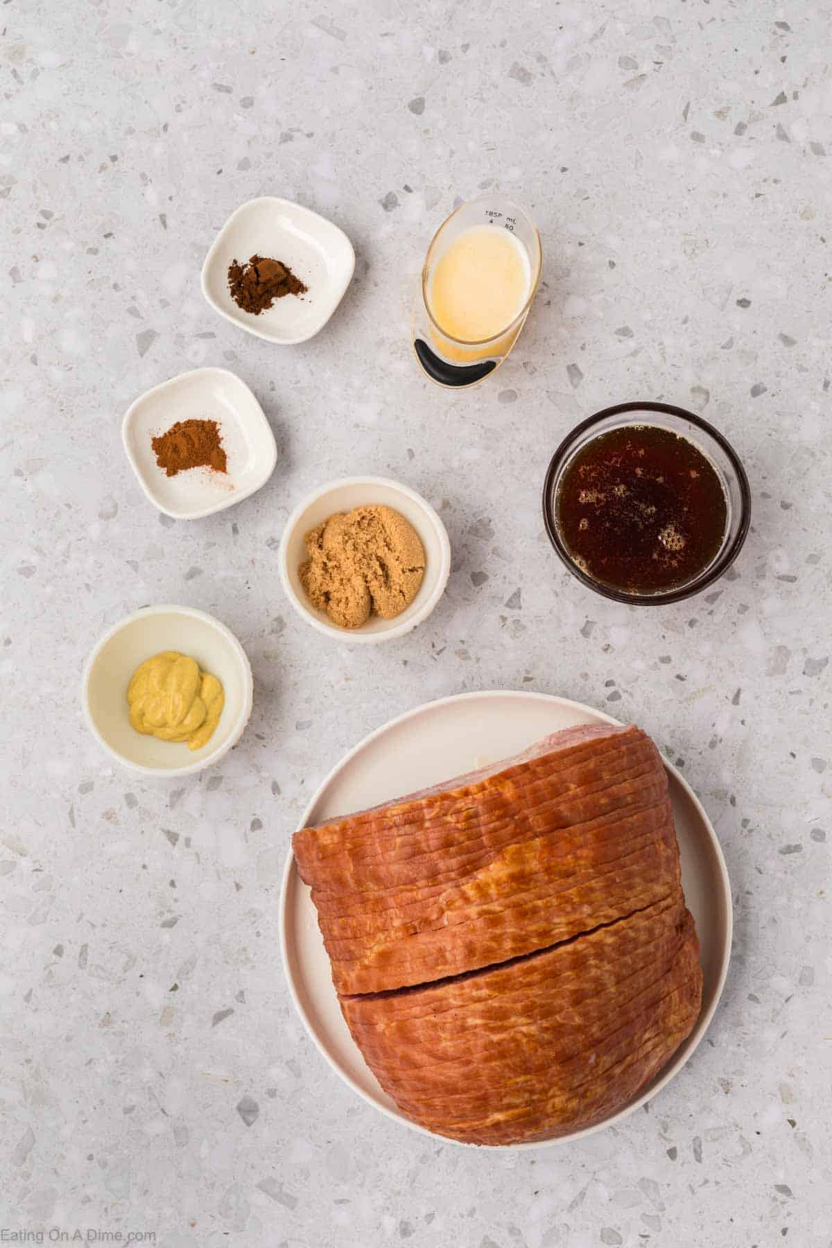 Un jamón en rodajas está ingeniosamente dispuesto en un plato blanco, rodeado de pequeños tazones de azúcar moreno, mostaza, especias molidas y un vaso de líquido oscuro sobre la encimera moteada, perfecto para un festín con freidora.