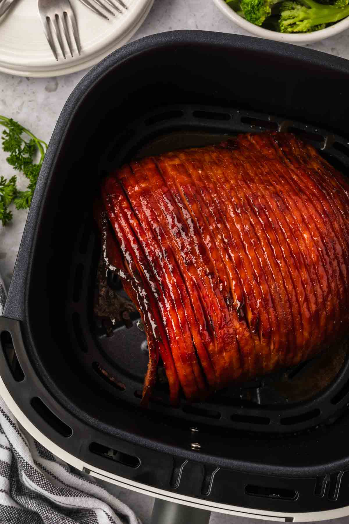 Un delicioso jamón glaseado chisporrotea a la perfección en la canasta de la freidora, con su superficie caramelizada y tentadora. Al fondo aguardan tenedores y platos, acompañados de una ramita de perejil fresco sobre una superficie de color claro.
