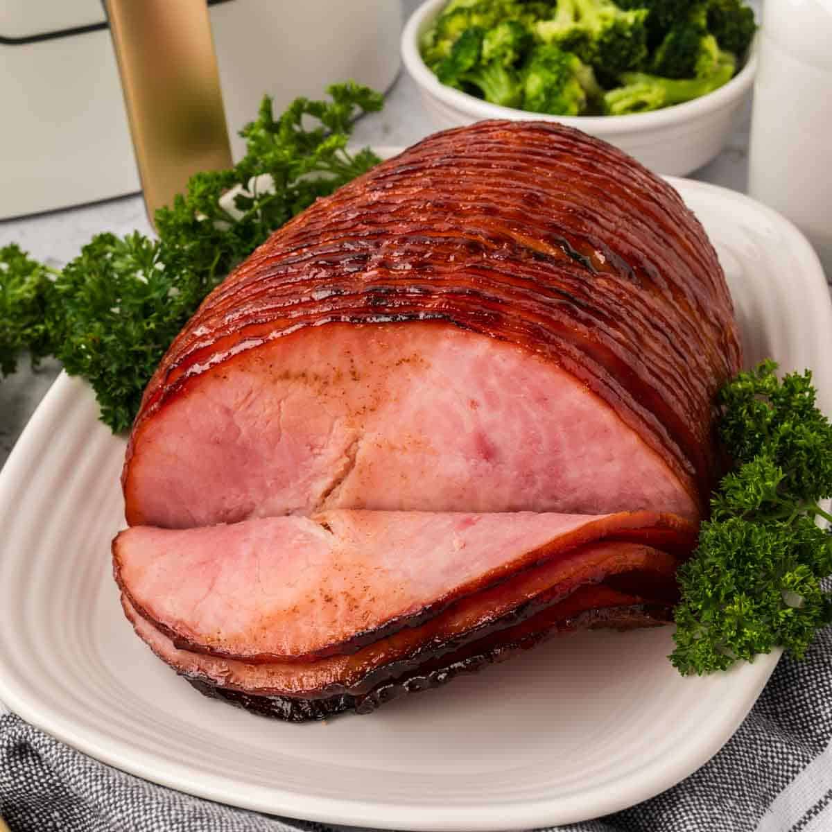 En una fuente blanca se muestra un jamón glaseado para freidora con algunas rodajas cortadas. El jamón se adorna con perejil y se acompaña de un plato de brócoli de fondo.