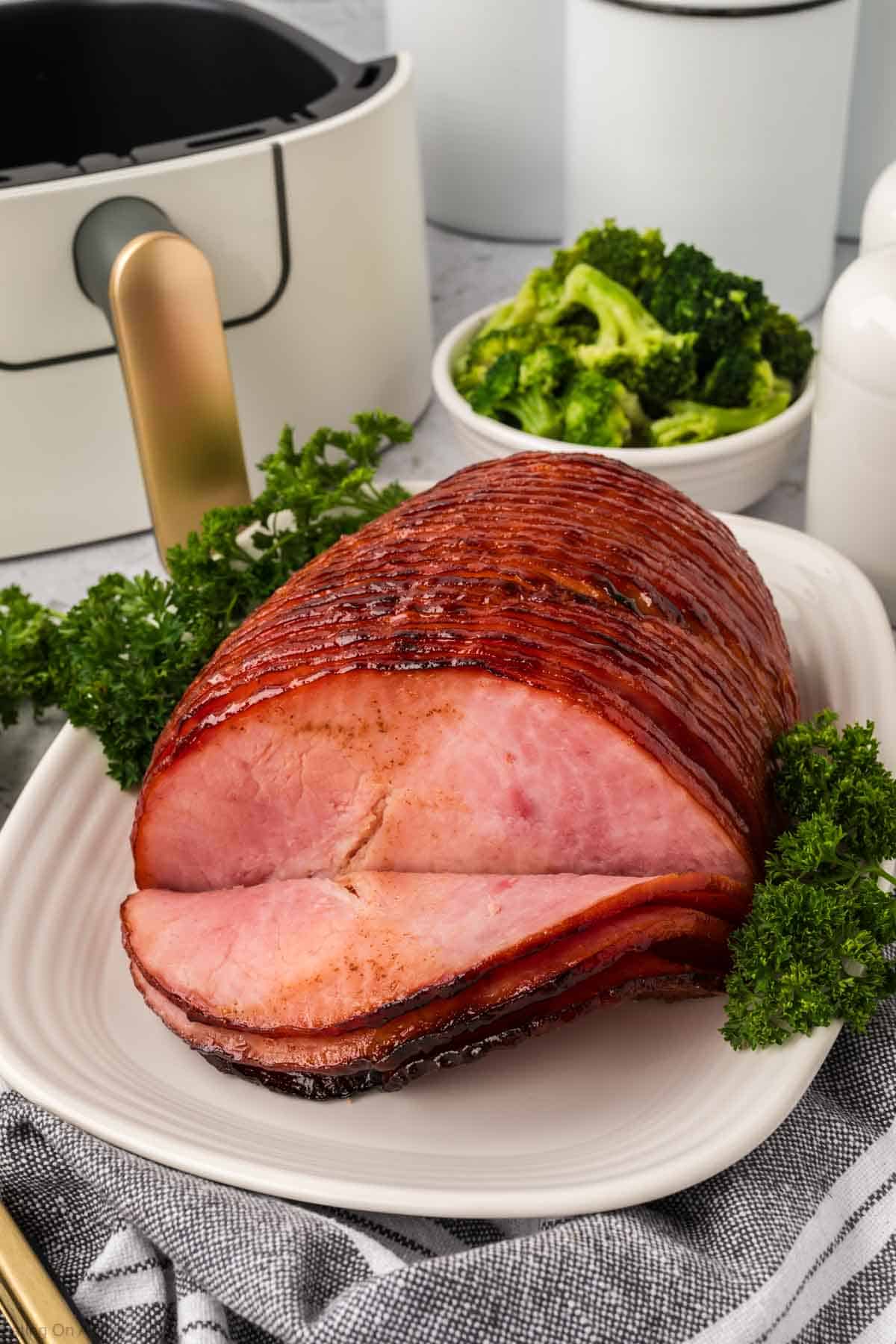 Un suculento jamón glaseado con freidora descansa sobre un plato blanco adornado con perejil. Los trozos de brócoli aparecen en un pequeño cuenco blanco al fondo. Un utensilio con mango dorado y un paño a rayas completan el atractivo paisaje de la mesa junto al plato.