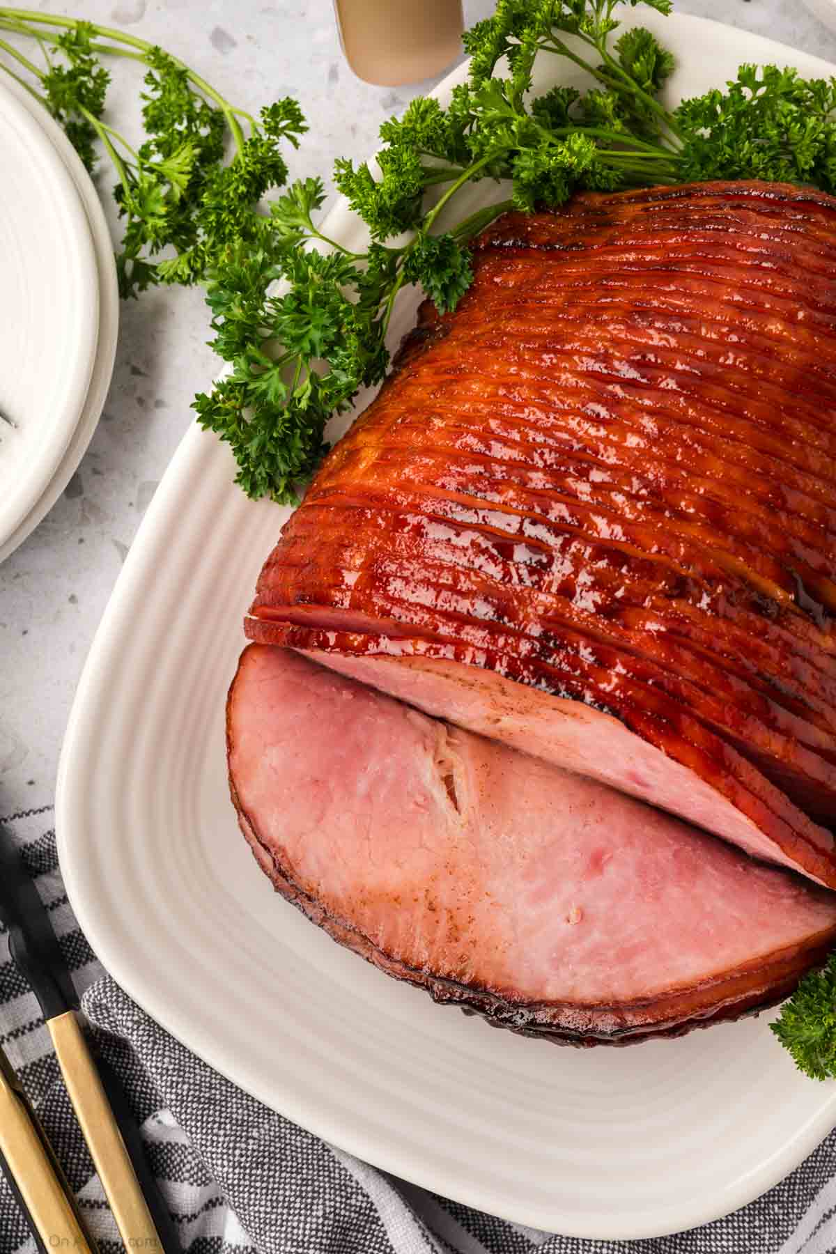 Un jamón glaseado en rodajas en espiral sobre un plato blanco, adornado con perejil fresco, brilla con una apariencia caramelizada. Cocinado a la perfección en una freidora, se asienta de manera atractiva sobre una encimera liviana junto a un paño a rayas y utensilios.