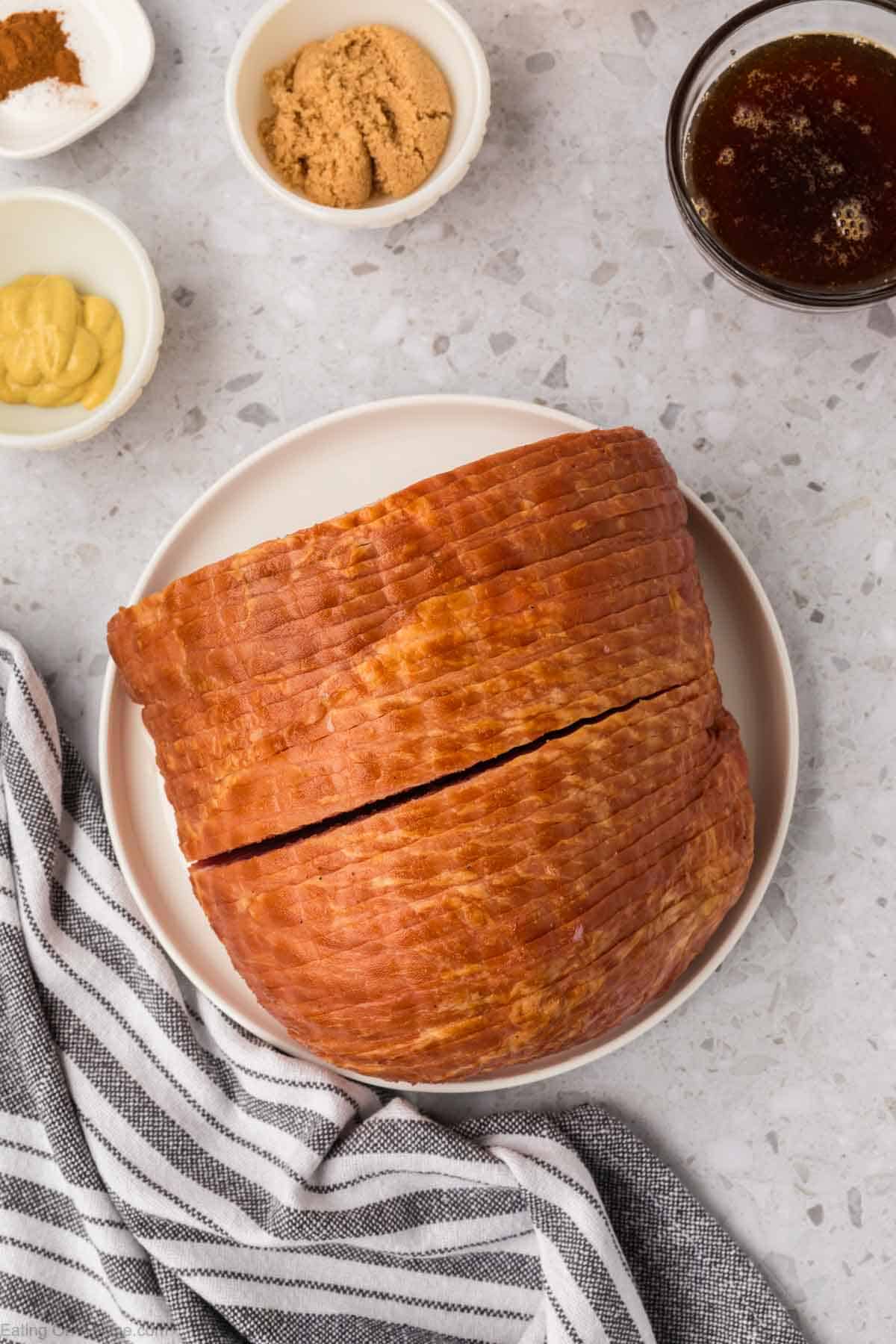 Sobre un plato blanco hay un jamón glaseado, rodeado de pequeños tazones de azúcar moreno, mostaza y especias sobre una encimera moteada. Perfecta para una receta fácil, una freidora puede simplificar la preparación de este plato. Cerca hay una servilleta de tela a rayas.