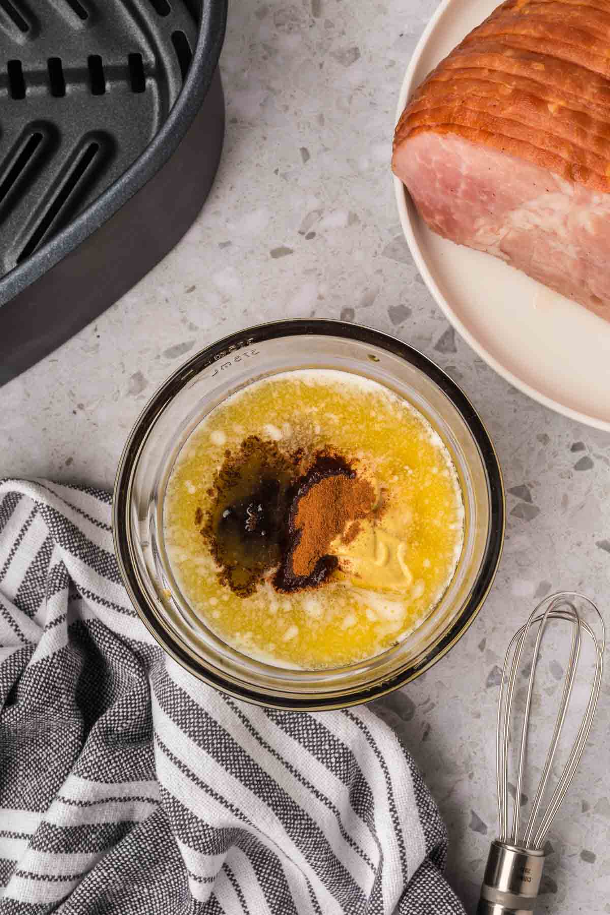 Un recipiente de vidrio sobre la encimera contiene mantequilla derretida con canela y azúcar moreno. Cerca, una freidora fríe un jamón cortado en espiral en un plato, mientras se ven un batidor, un paño rayado y el mango de una fuente para asar.