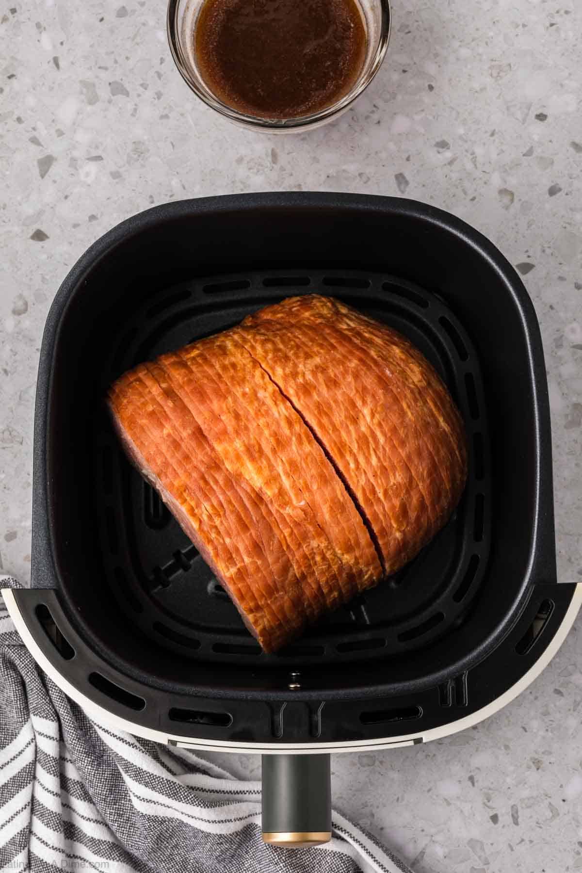 Un jamón rebanado descansa dentro de la canasta de una freidora sobre una encimera moteada. Cerca, espera un cuenco de glaseado marrón, mientras que un paño de cocina a rayas se asoma desde la parte inferior izquierda.