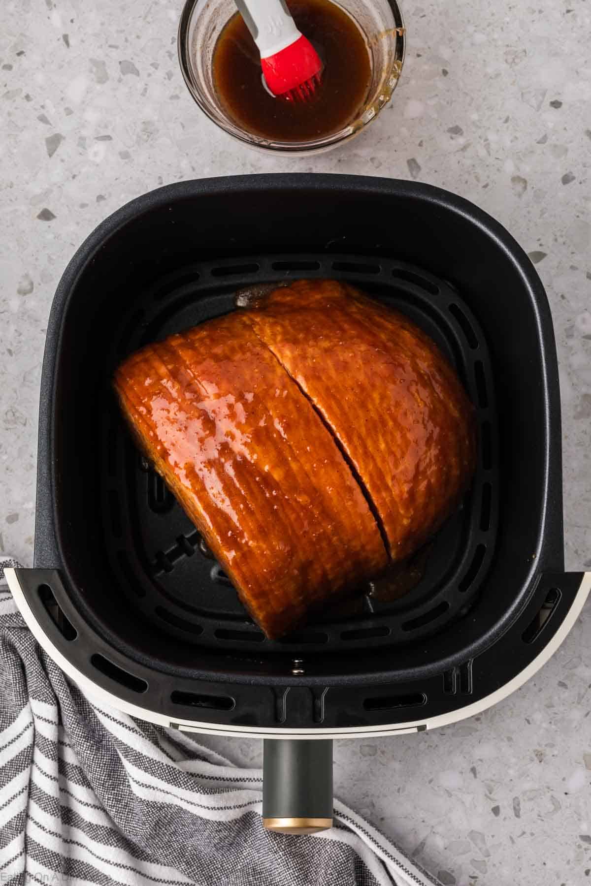 Un delicioso jamón glaseado chisporrotea en la freidora, con una porción perfectamente cortada. Un pequeño cuenco de esmalte y un pincel se encuentran en la encimera moteada al lado, mientras que una tela a rayas blancas y negras se asoma a la vista en la parte inferior de la imagen.