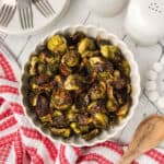 A white scalloped bowl filled with Balsamic Brussels Sprouts sits on a white wooden table, next to a red and white towel, a wooden spoon, salt and pepper shakers, and stacked plates with forks.