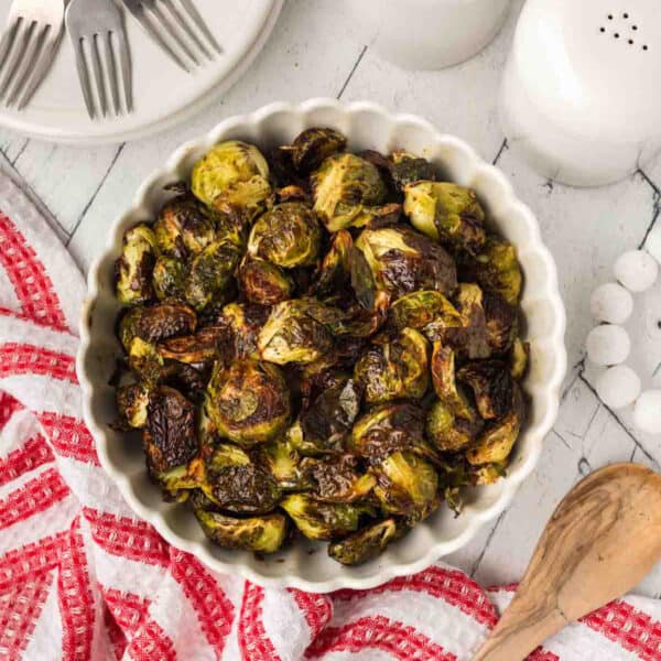 A white scalloped bowl filled with Balsamic Brussels Sprouts sits on a white wooden table, next to a red and white towel, a wooden spoon, salt and pepper shakers, and stacked plates with forks.