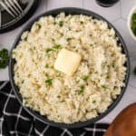 A bowl of butter rice, topped with a pat of butter and sprinkled with chopped parsley, sits on a table beside a black and white checkered napkin.