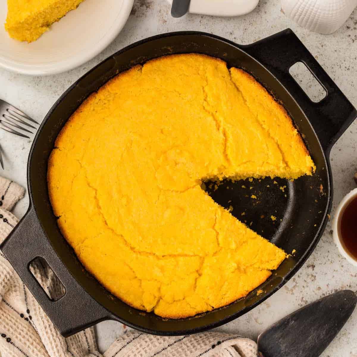 Una bandeja redonda de pan de maíz dorado al que le falta una rebanada se encuentra atractivamente en una sartén de hierro fundido negra sobre una superficie de color claro. Cerca hay utensilios, una toalla beige y un plato blanco listo para acompañar tu delicia de café matutino.