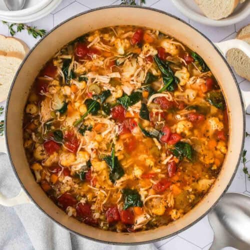 A large pot filled with hearty leftover turkey soup and vegetables, featuring chunks of tomato, shredded turkey, carrots, spinach, and herbs in a clear broth. Slices of bread are visible on the side.