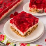A square slice of Raspberry Pretzel Salad with a crumbly base, creamy middle layer, and bright red raspberry gelatin topping is served on a white plate, garnished with fresh raspberries and another serving in the background.
