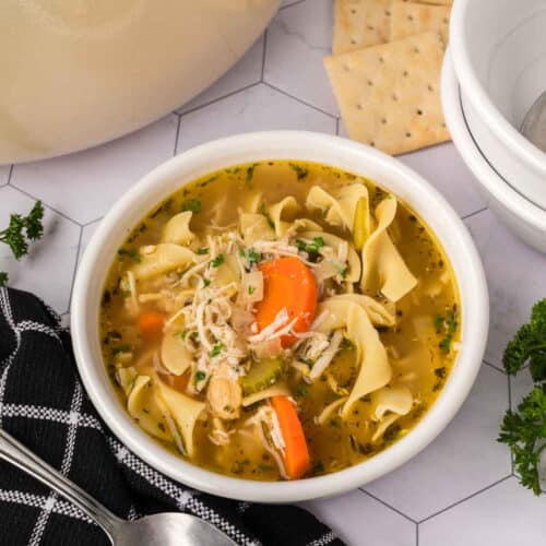 A bowl of Turkey Noodle Soup with carrots, celery, and egg noodles sits on a white surface beside a black and white napkin, a spoon, parsley garnish, crackers, and stacked empty bowls.