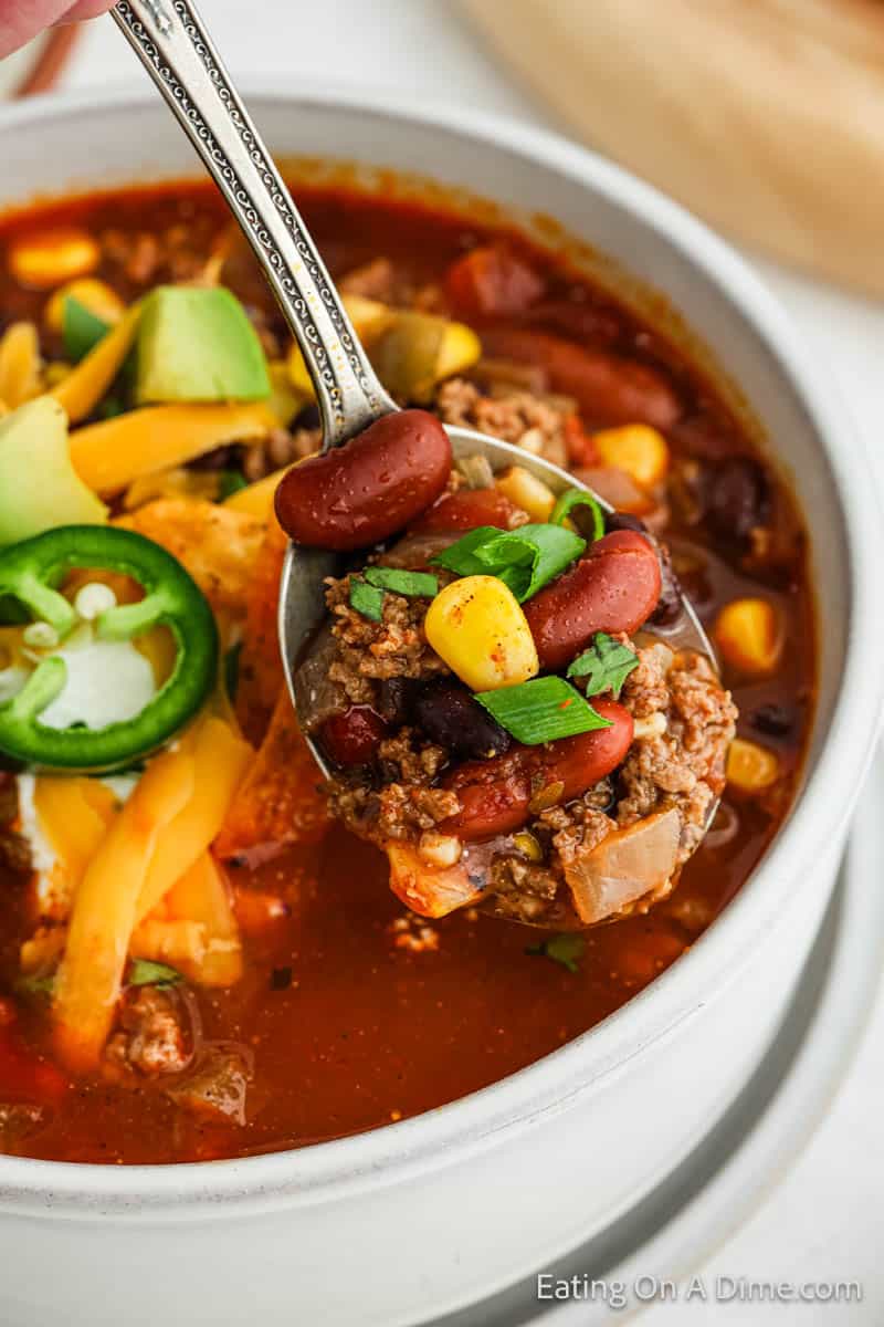 A spoonful of chili, reminiscent of a hearty taco soup, with ground beef, red kidney beans, corn, and diced vegetables is held above a bowl. The bowl brims with chili and is garnished with sliced jalapeño, shredded cheese, avocado, and sour cream—a delectable recipe to savor.