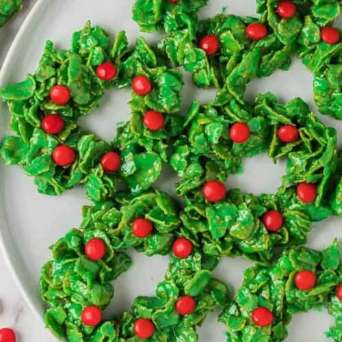 Green cornflake Christmas Wreath Cookies, decorated with small red candy pieces, are artfully arranged on a white plate.