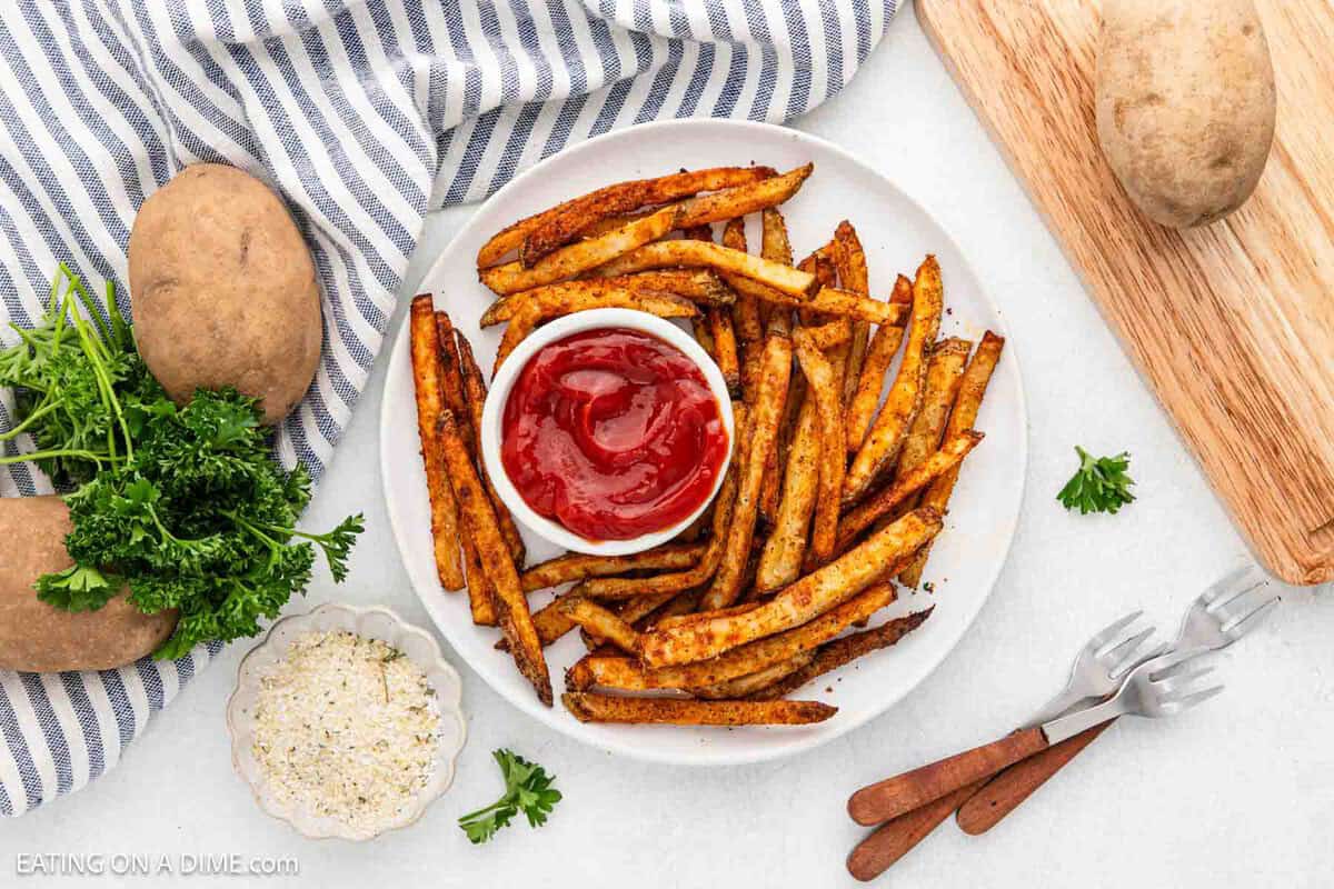 A plate of homemade, baked French fries seasoned with spices sits invitingly beside a small dish of ketchup. Surrounding the plate are raw potatoes, a striped cloth, fresh parsley, and a bowl of seasoning. Two forks rest nearby on the pristine white surface.