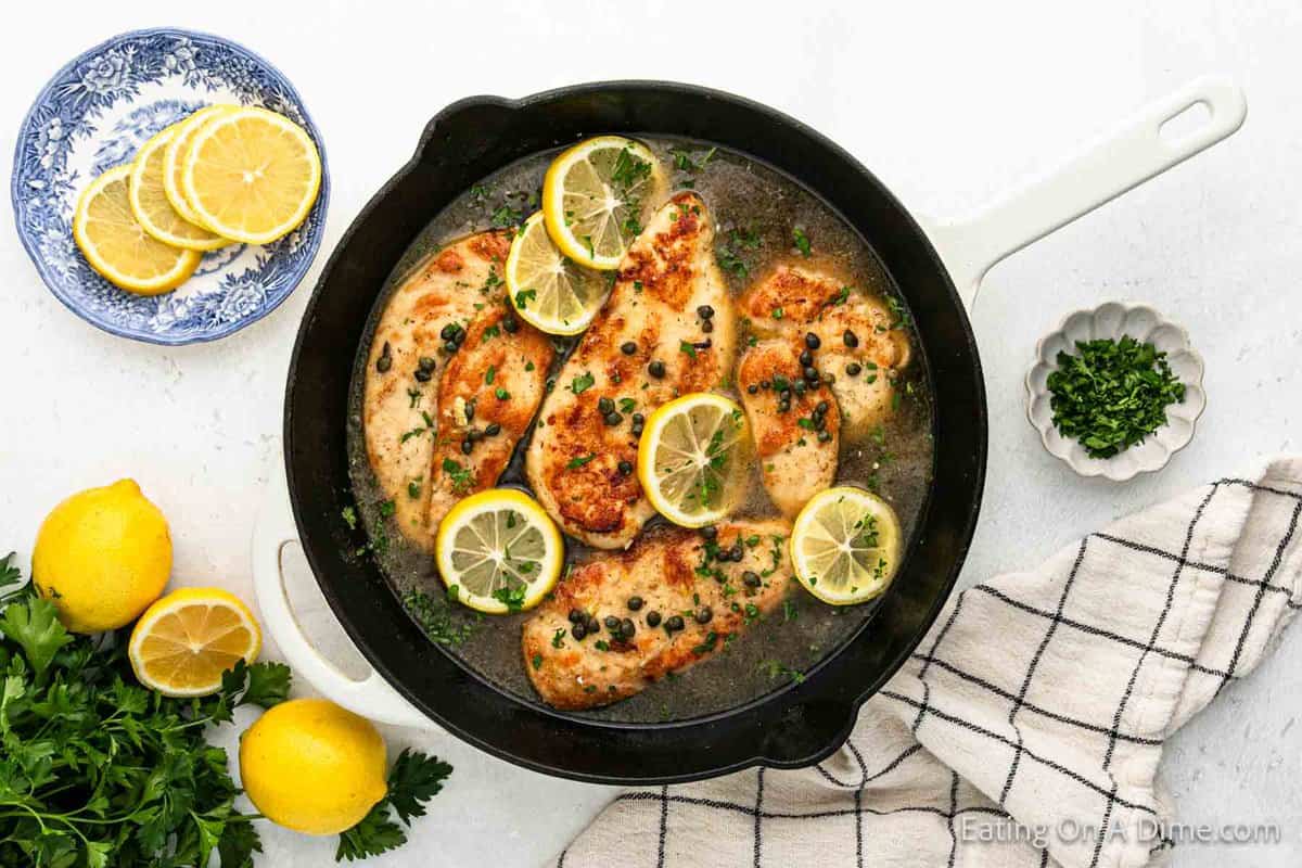 A skillet filled with Chicken Piccata, an Italian dish of cooked chicken breasts garnished with lemon slices and capers, rests on a white surface.