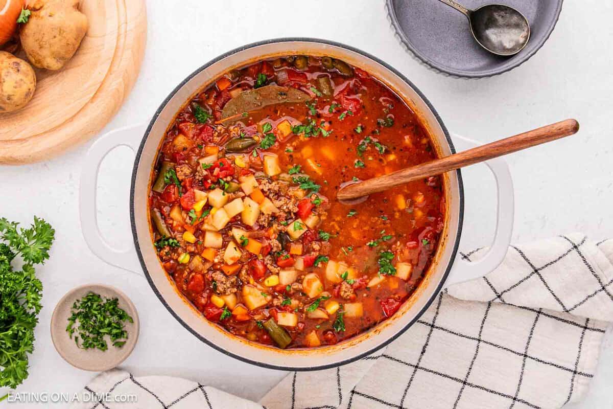 A pot of hearty vegetable soup, reminiscent of a classic hamburger soup, brims with diced potatoes, green beans, tomatoes, and herbs. A wooden spoon rests inside on a light countertop with a checkered cloth nearby. A small bowl of chopped herbs and potatoes are on the side for the perfect recipe touch.