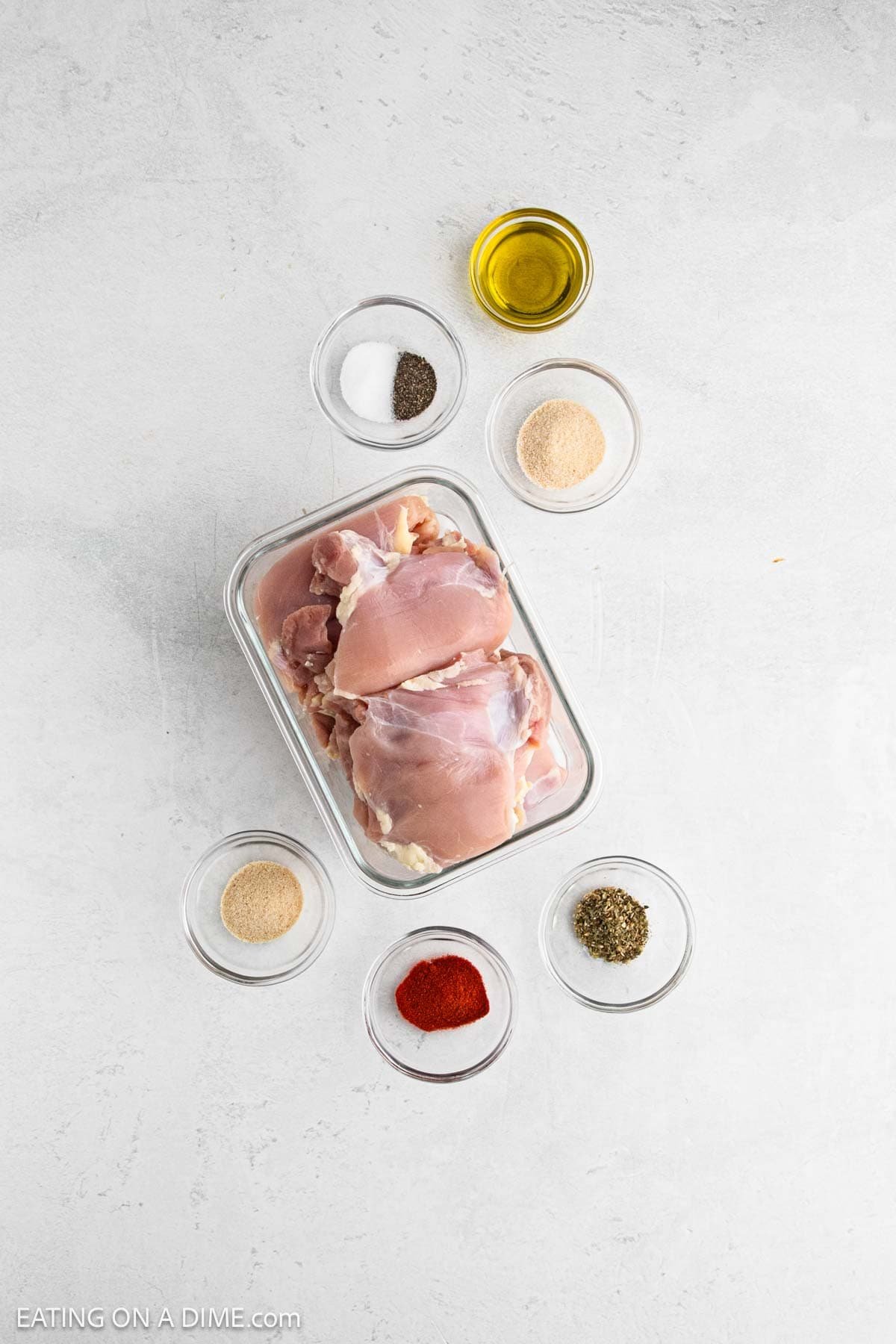 Raw boneless chicken thighs in a clear glass dish, surrounded by small bowls of olive oil, spices, and Italian seasoning—perfect ingredients for delicious Air Fryer Chicken Thighs on a light gray surface.