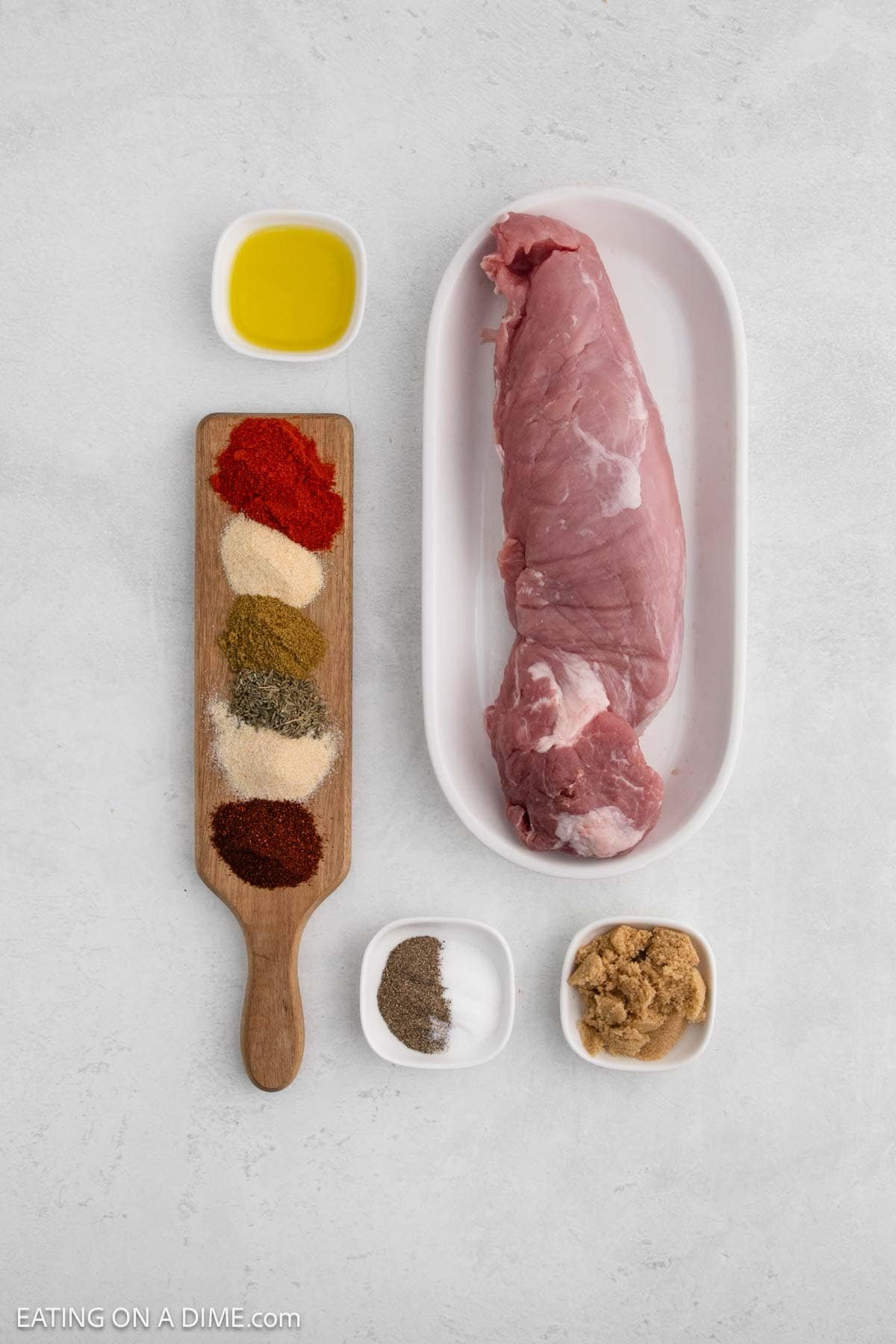 Raw pork tenderloin on a white tray, ready for the air fryer, surrounded by small bowls of olive oil, brown sugar, salt, pepper, and a wooden board with piles of various spices on a light-colored surface.