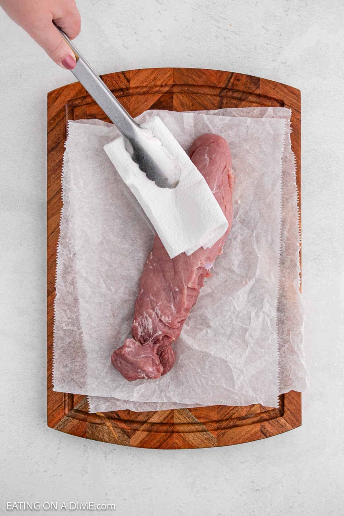 A hand uses tongs and a paper towel to pat dry a raw pork tenderloin on a wooden cutting board lined with parchment paper, preparing it for air fryer pork tenderloin.